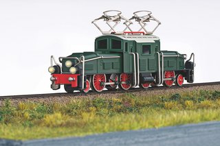 Modell einer grünen Elektrolokomotive mit roten Rädern auf Gleisen, im Vordergrund Wiese und Begrünung.