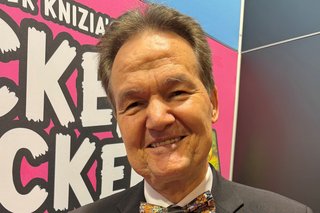 Reiner Knizia is standing in front of an exhibitor with the sign "Mücken Drücken".