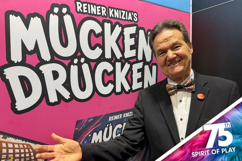  Reiner Knizia steht vor einem Austeller mit der Aufschrift ,,Mücken Drücken" 