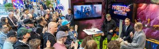 Viele bunte Maskottchen und Menschen posieren gemeinsam in einer Messehalle.