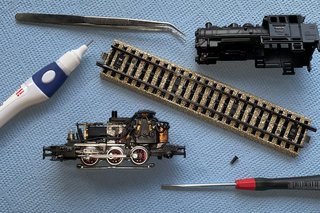 Zerlegte Modelllokomotive mit Werkzeug, Gleisstück und Kleinteilen auf einer blauen Unterlage.