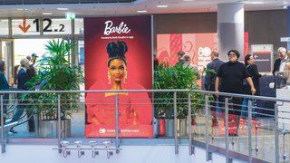 Large Barbie ad poster with red background, visitors and plants in exhibition area.