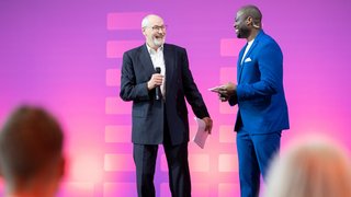 Two men talking on stage, one holding a microphone, purple background.