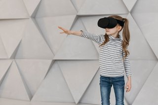 Kind vor grauen Hintergrund mit VR Brille auf und nach vorne zeigend