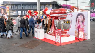 Bunte Playmobil-Werbeplakate vor Messehalle, Besucher gehen daran vorbei.
