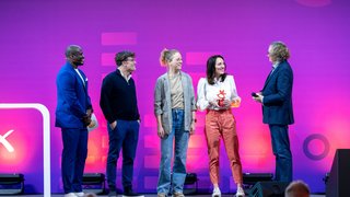 Five people stand on stage, one holds a trophy, purple background.