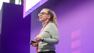 Woman with microphone gives a talk on stage with purple background at Toy Business Forum.