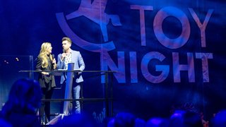Two hosts speak on stage in front of large "TOY NIGHT" lettering.