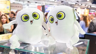 Two white plush figures with big yellow eyes sit on a glass table at the fair.