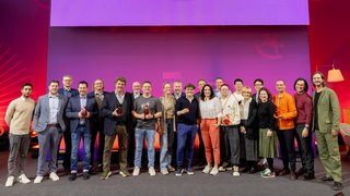 Group of award winners stands with trophies on stage in front of a purple background.