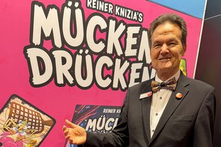 Reiner Knizia is standing in front of an exhibitor with the sign "Mücken Drücken".