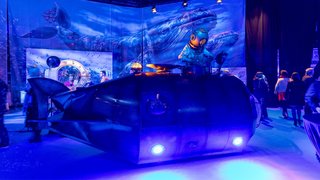 Submarine set with diver figure, blue whale in background, visitors in exhibition room.