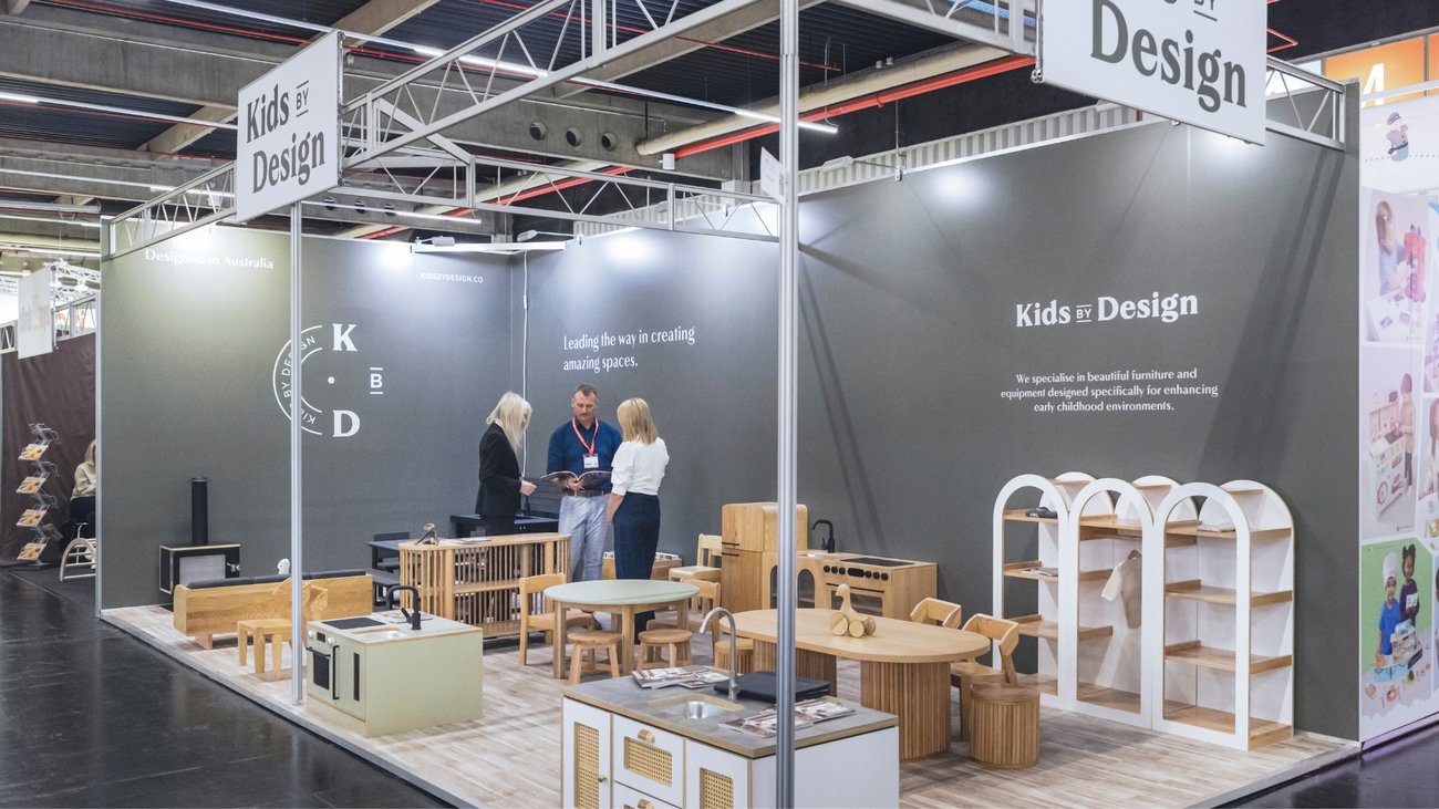 Messestand mit Holz-Kindermöbeln und Regalen, drei Personen im Gespräch, Kids by Design.