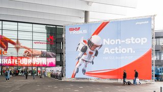 Menschen laufen an großem Magna-Tiles-Werbeplakat vor Messegebäude vorbei.